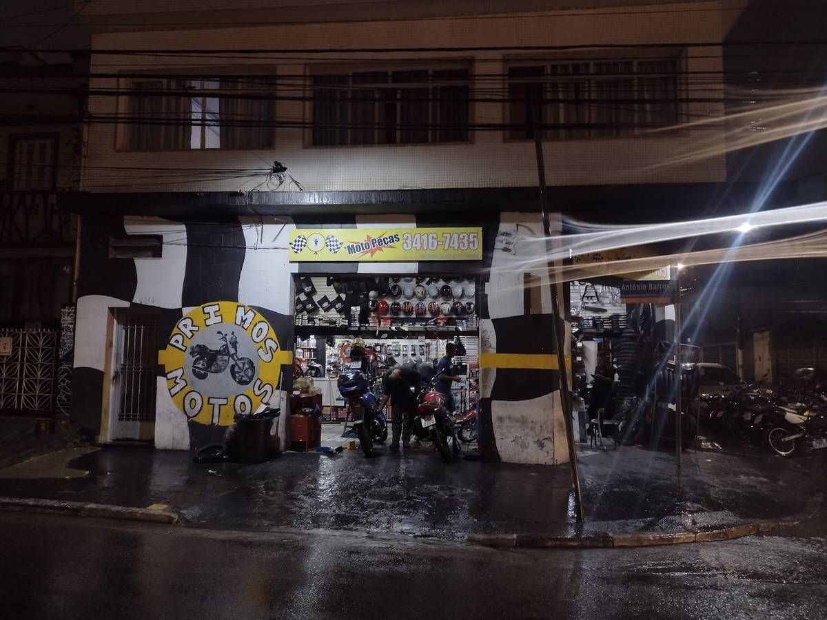 Fachada da Primos Motos à noite, com o muro pintado de bandeira quadriculada, capacetes pendurados na entrada e duas motos paradas dentro da loja.