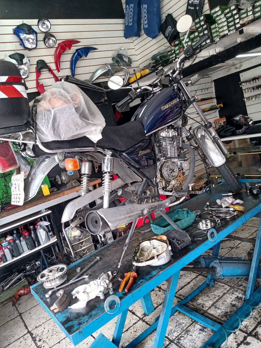 Moto Suzuki com o motor aberto sobre a bancada azul da oficina, com peças, escapamento e ferramentas espalhados ao lado.